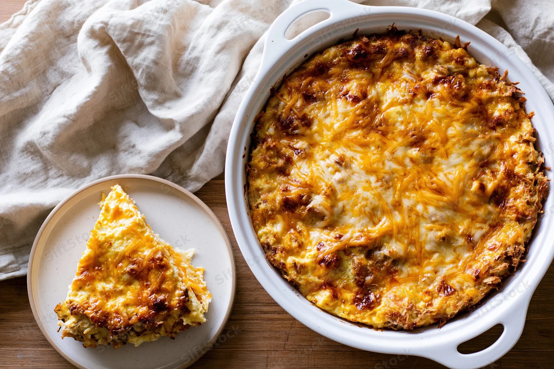 Golden-brown cheesy casserole in white ceramic dish with slice served on plate beside it on wooden table with cloth napkin