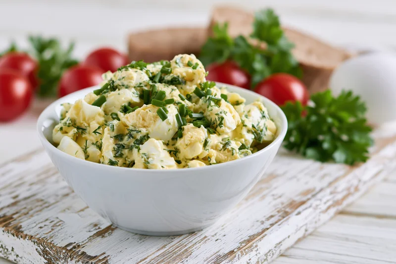 Chunky egg salad with chives and parsley in a white ceramic bowl on a rustic wooden surface