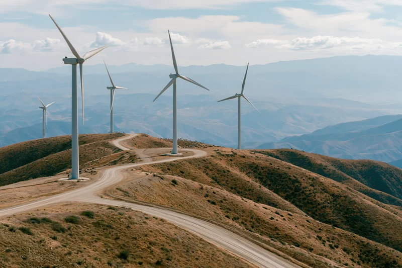 Wind turbines arranged across rolling arid hills with dirt road winding through the terrain under a partly cloudy sky