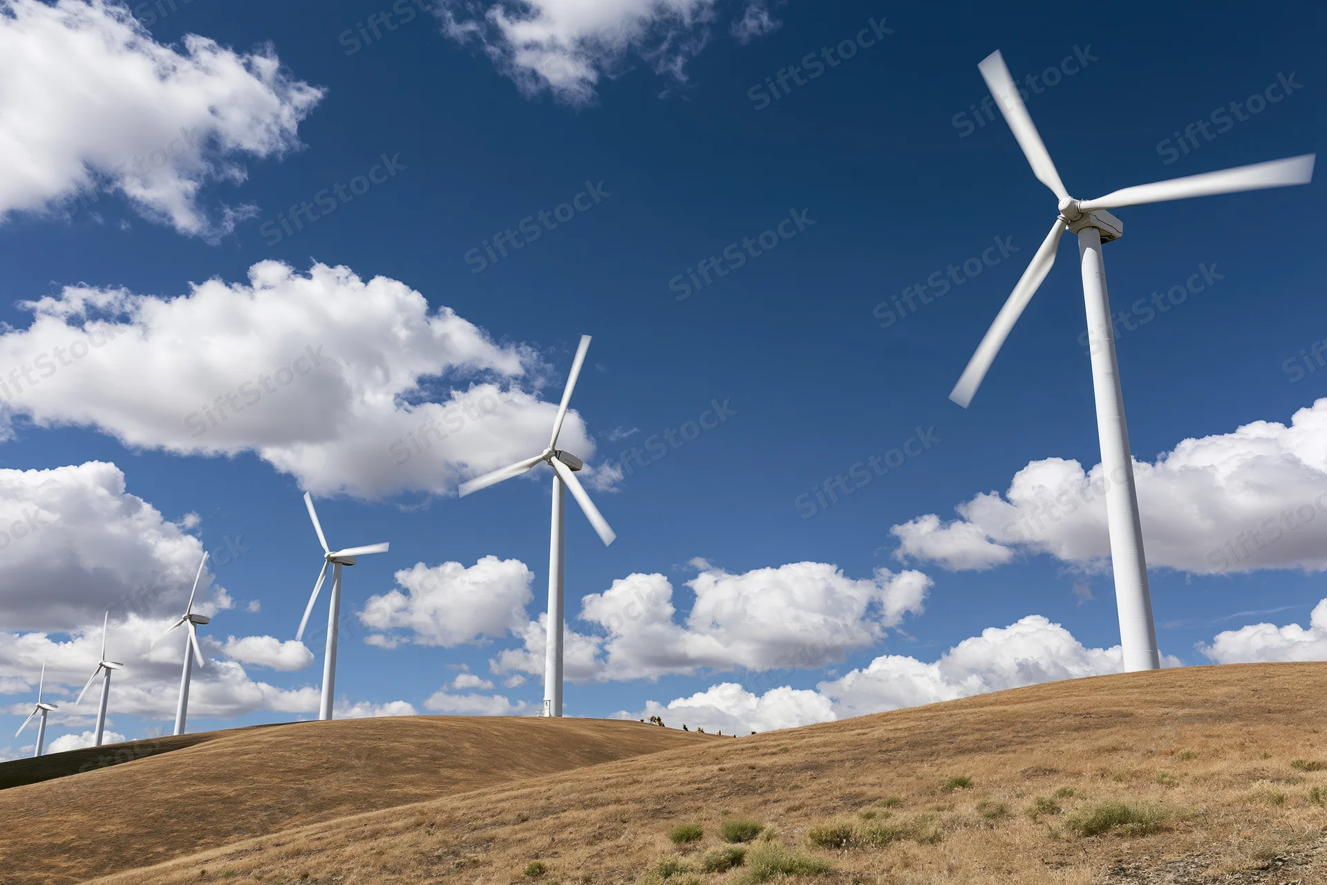 Wind turbines standing on rolling brown hills under a blue sky with white clouds