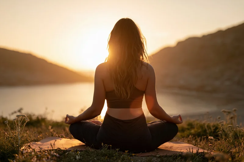 Woman meditating in lotus pose on grassy hill overlooking lake at sunset