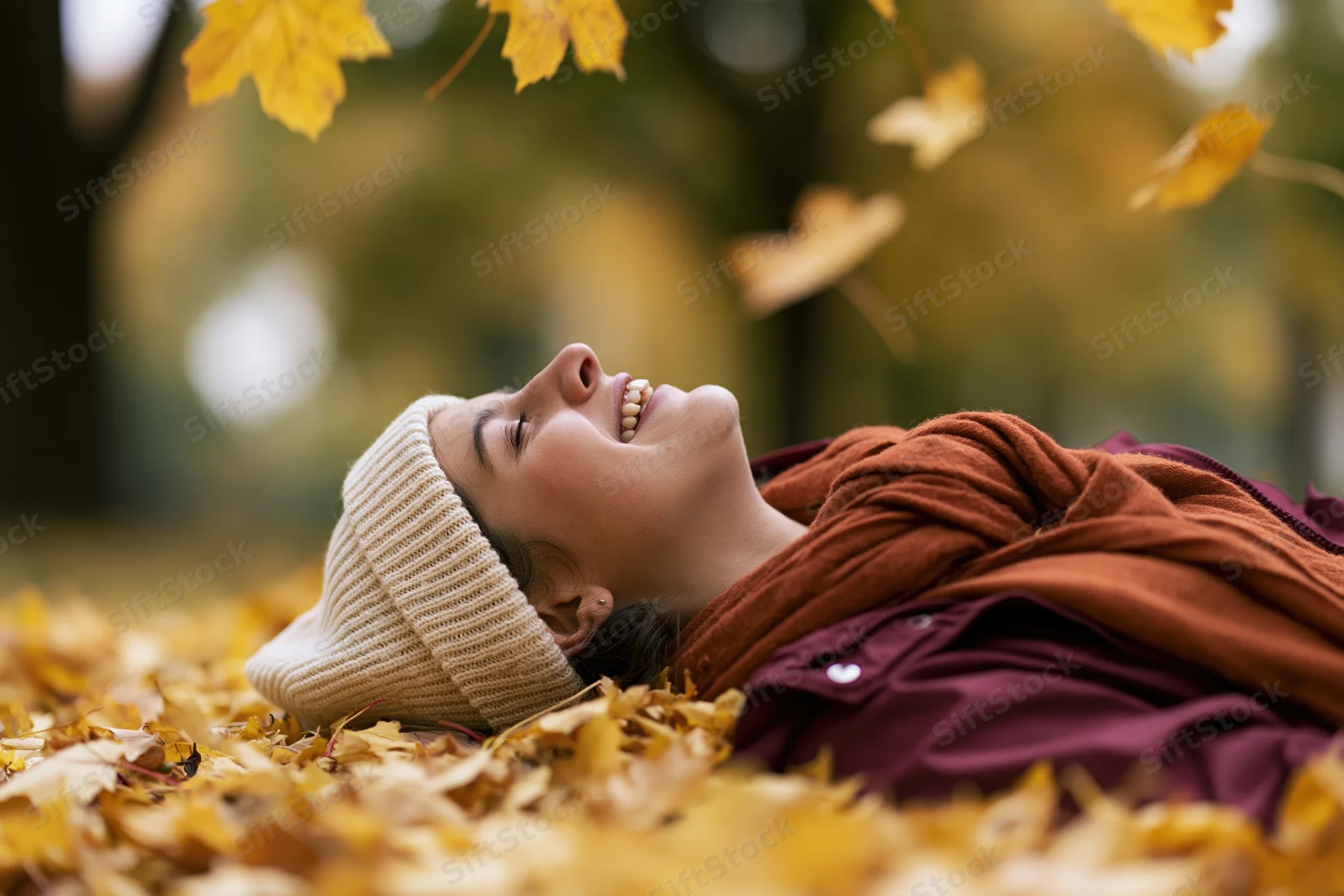 Young woman lying on back in pile of fallen autumn leaves smiling with eyes closed in park