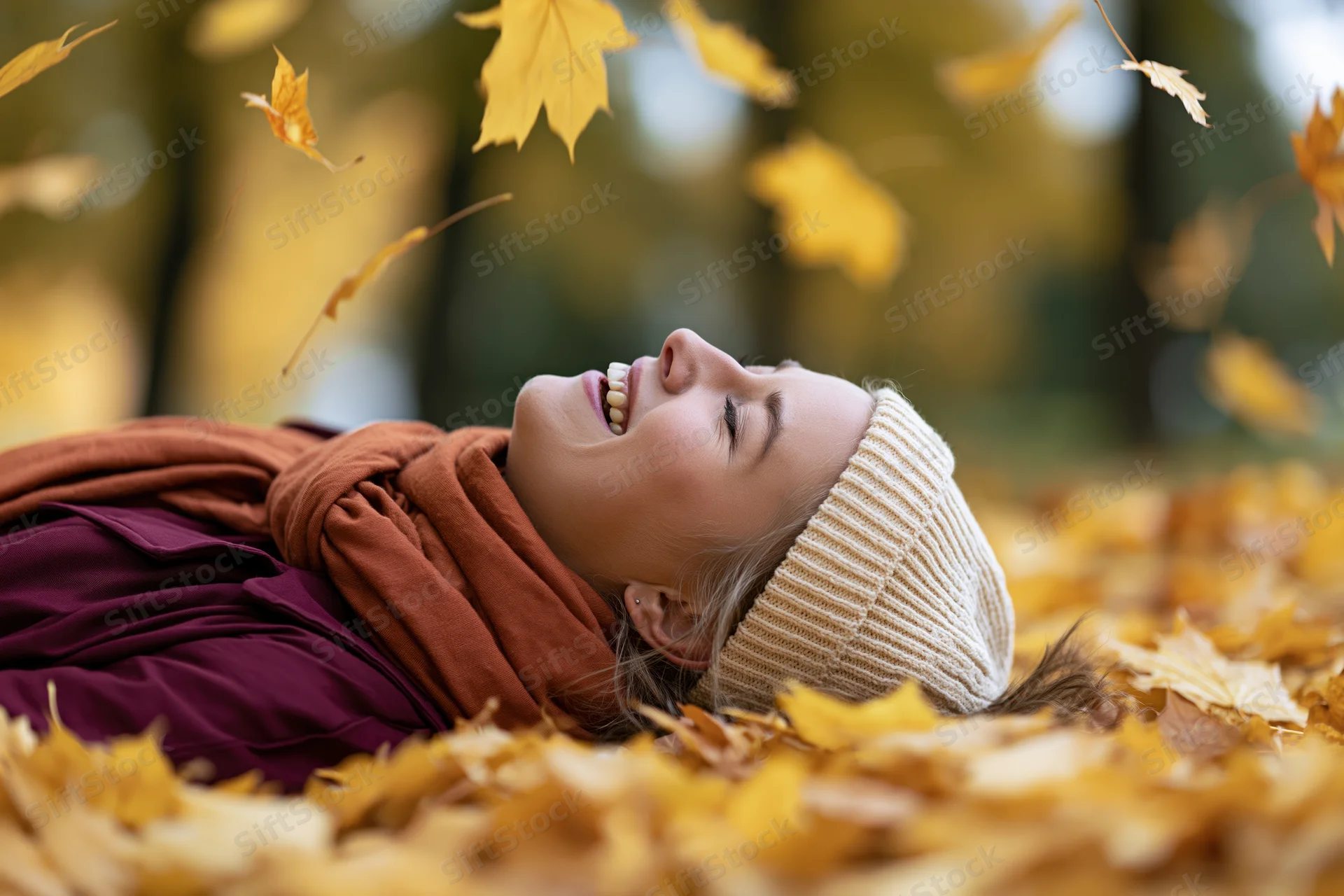 Young woman lying on back in pile of fallen autumn leaves smiling with eyes closed in park