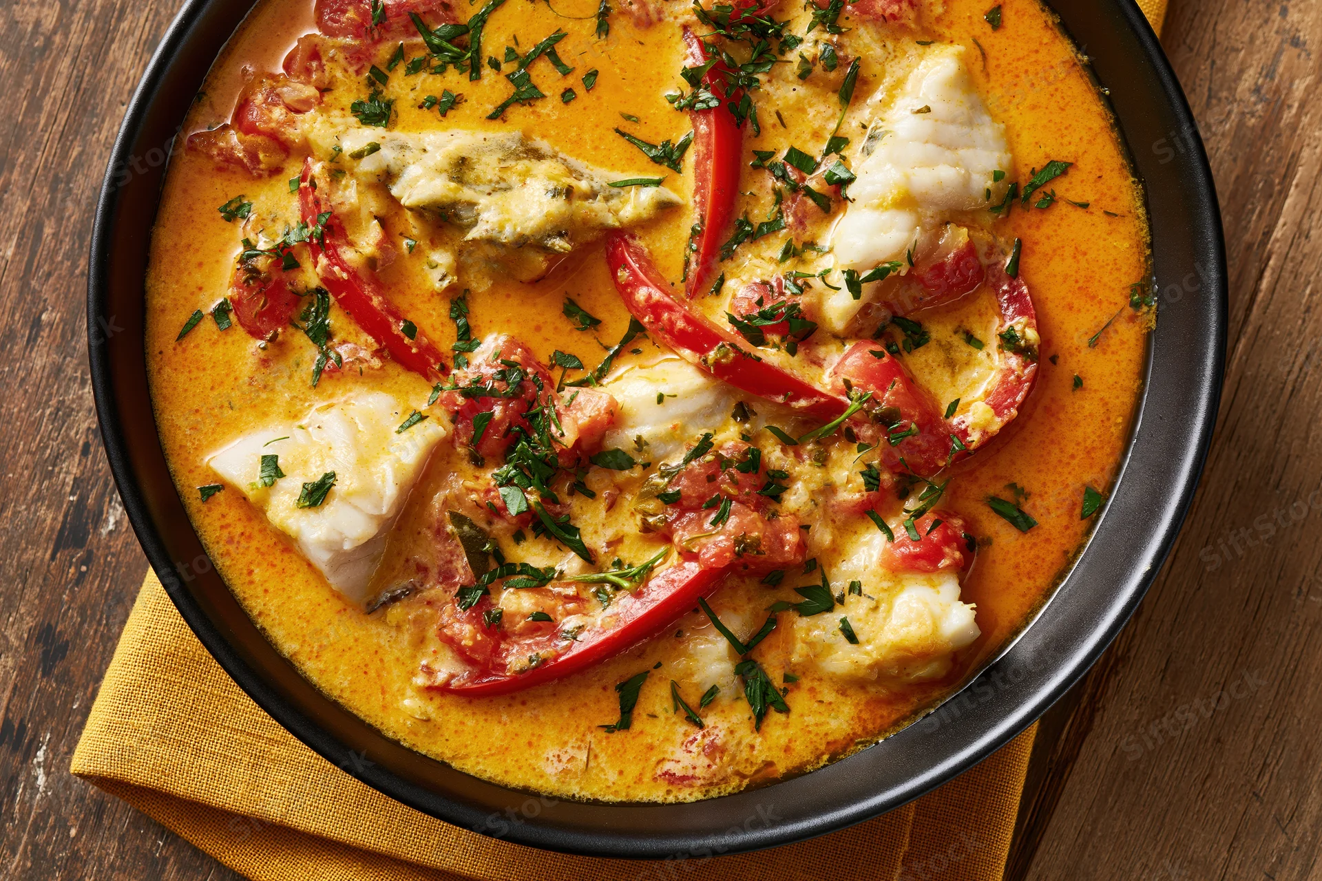 Creamy orange fish stew with tomatoes and peppers in a black bowl on a wooden table