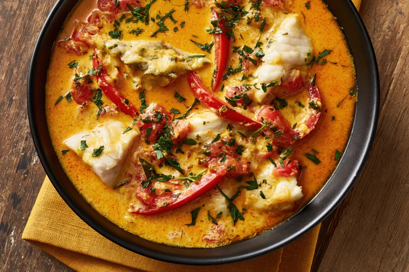 Creamy orange fish stew with tomatoes and peppers in a black bowl on a wooden table