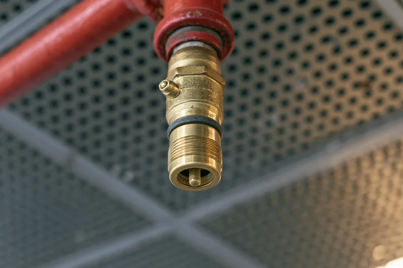 Brass fire sprinkler head mounted on red pipe against perforated ceiling