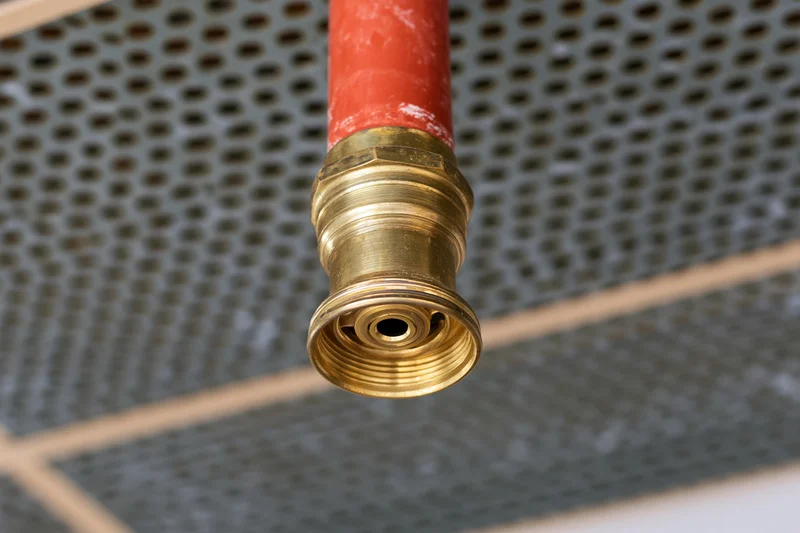 Brass fire sprinkler head mounted on red pipe against perforated ceiling grid