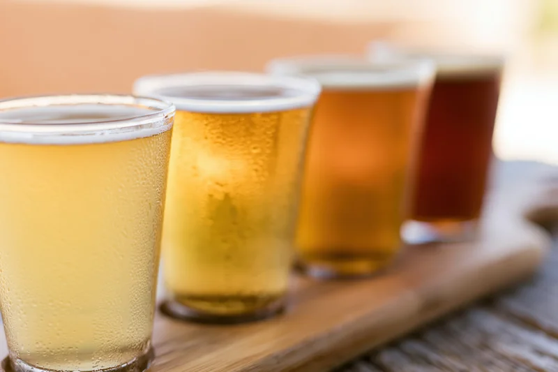 Four craft beers in small pint glasses arranged on a wooden tray with condensation and foam, showcasing variety in color and type
