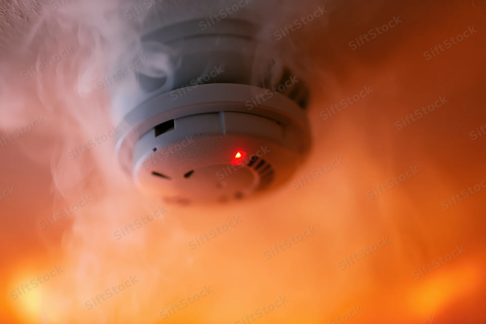Smoke detector mounted on ceiling with red light glowing amid rising smoke and orange flames