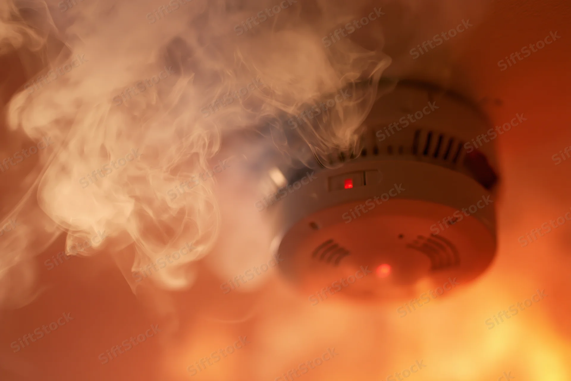 Smoke detector mounted on ceiling with smoke and red light activated during fire emergency