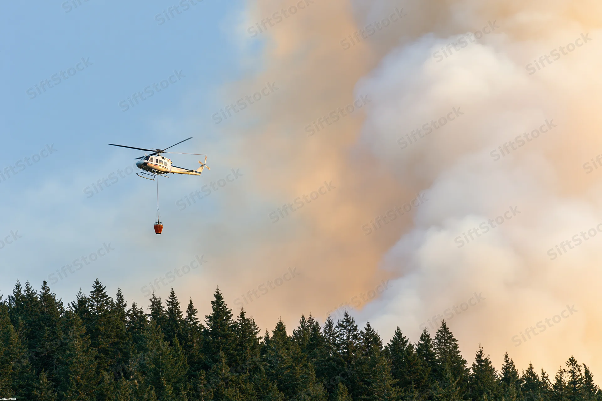 Helicopter carrying water bucket over forest with smoke rising