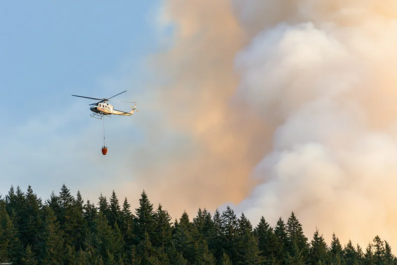 Helicopter carrying water bucket over forest with smoke rising