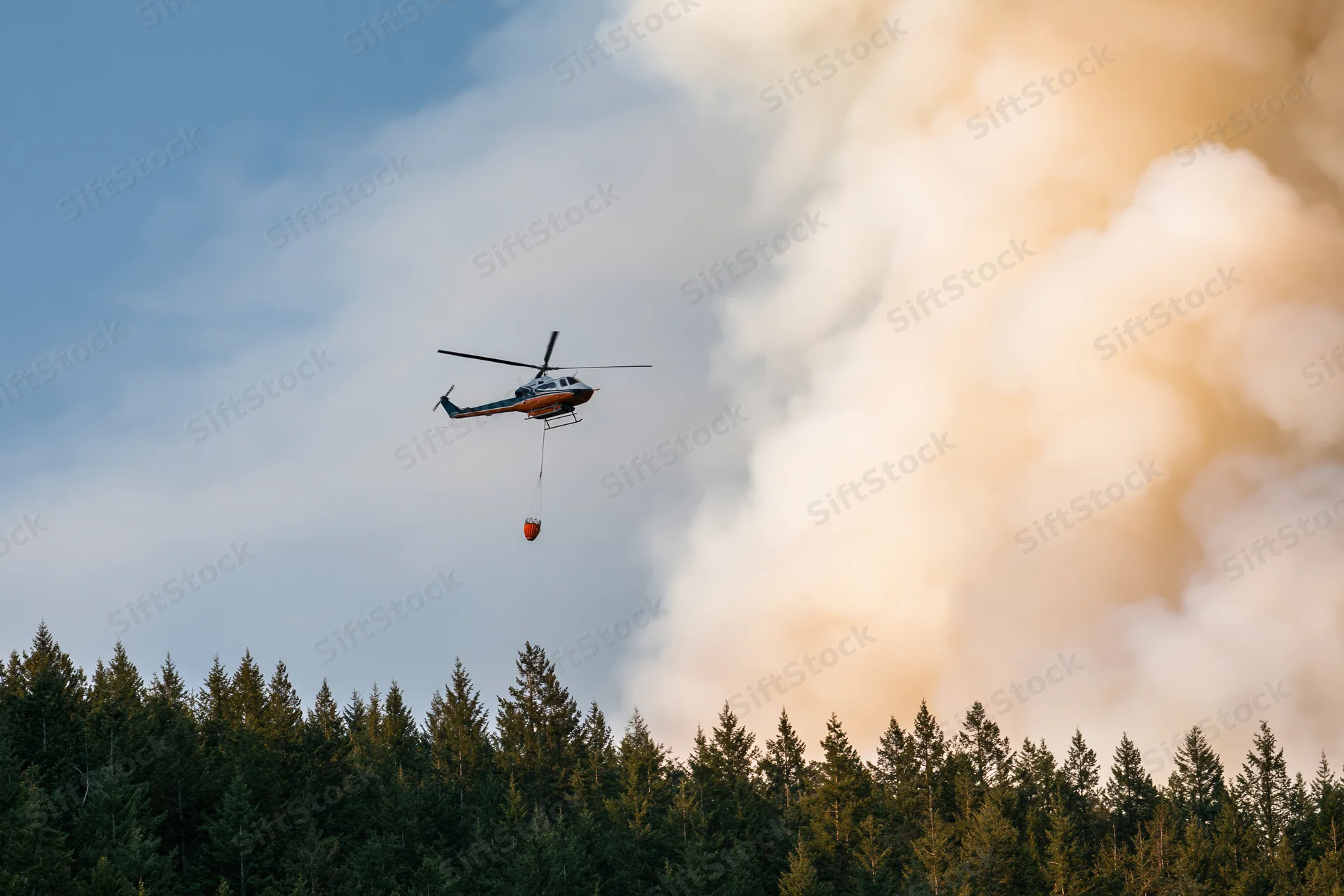Helicopter carrying water bucket over forest with smoke rising