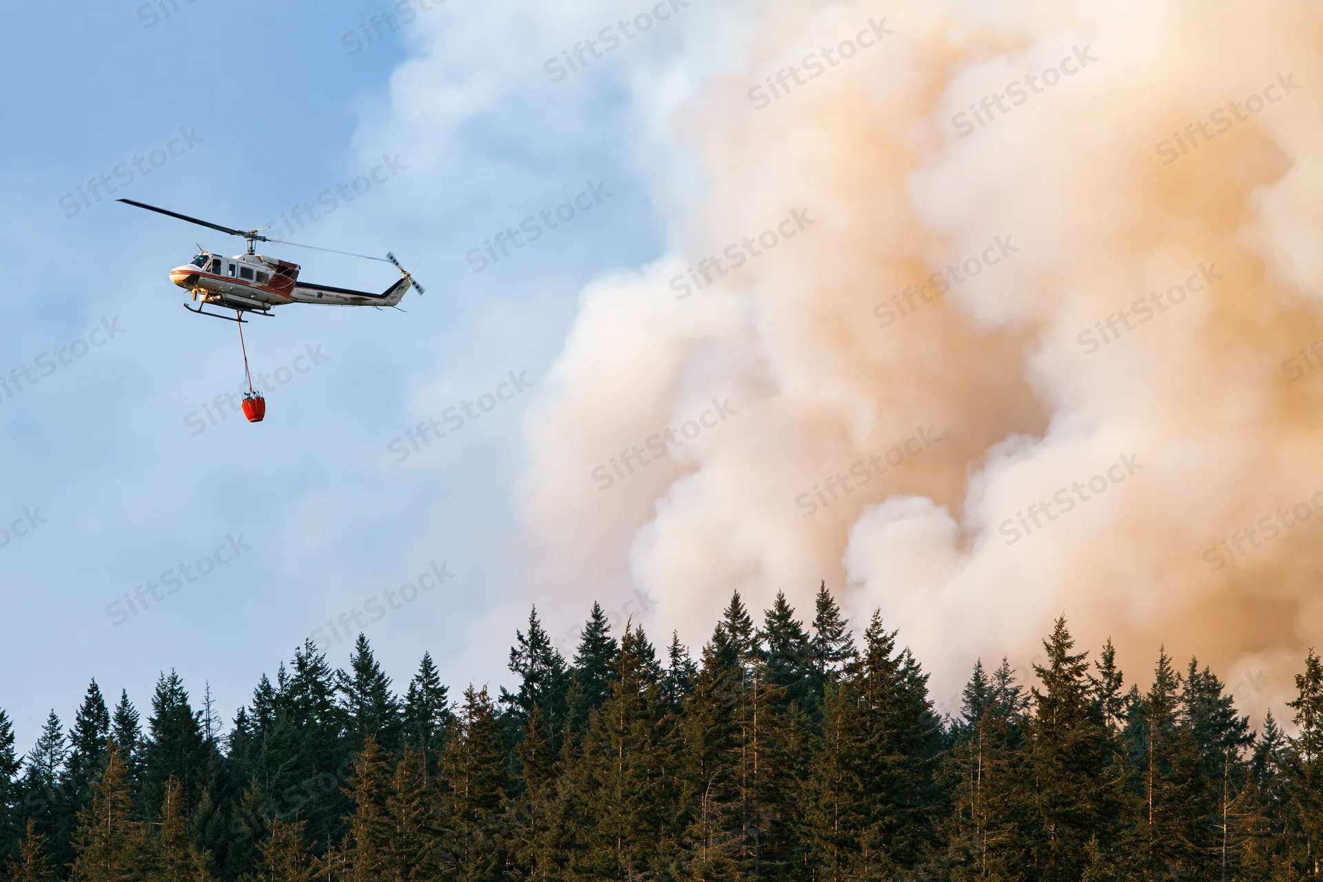 Helicopter carrying water bucket flying over forest with smoke rising from wildfire