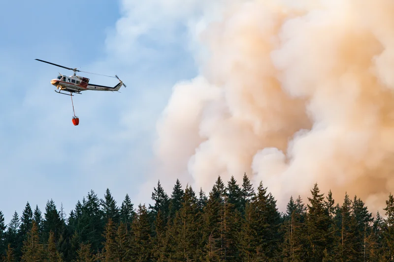 Helicopter carrying water bucket flying over forest with smoke rising from wildfire