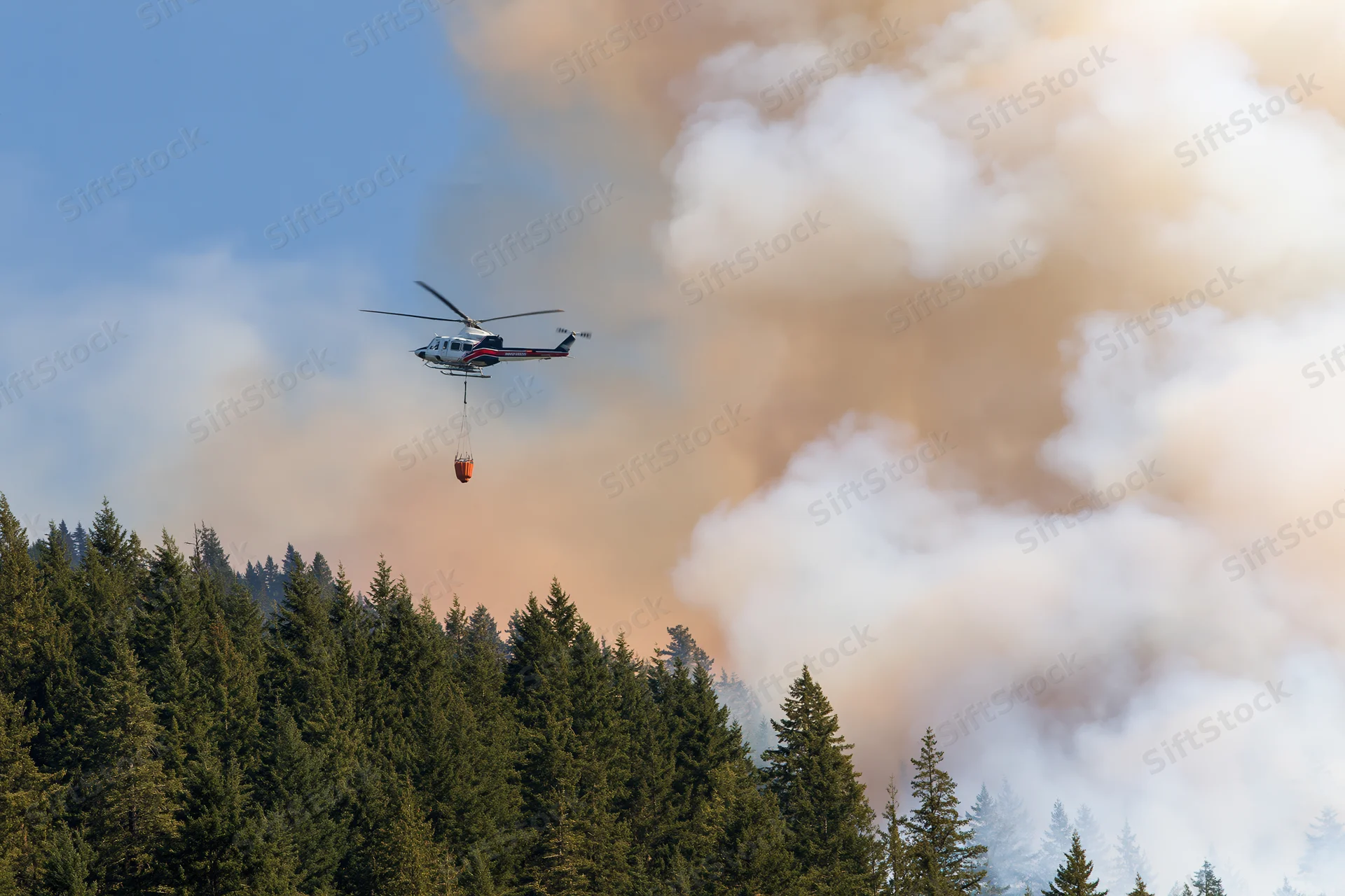 Helicopter carrying water bucket over forest during wildfire suppression operation