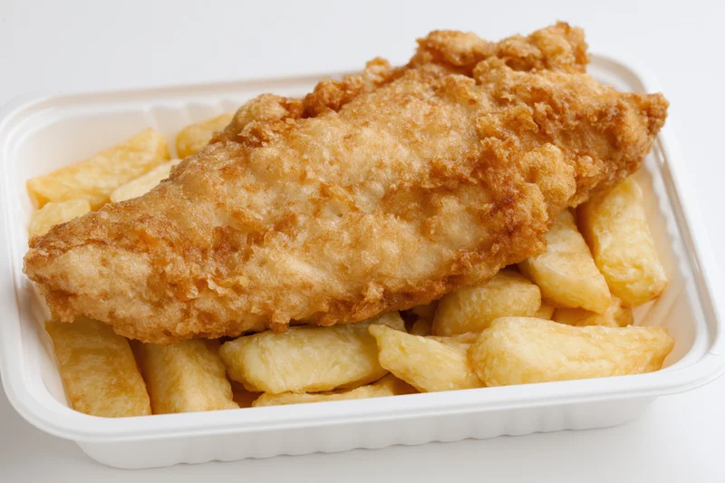Battered fish fillet resting on thick-cut chips in a white takeaway container