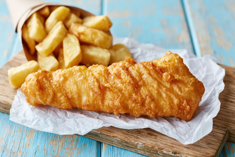 Golden battered fish fillet on crumpled white paper with chips on wooden board