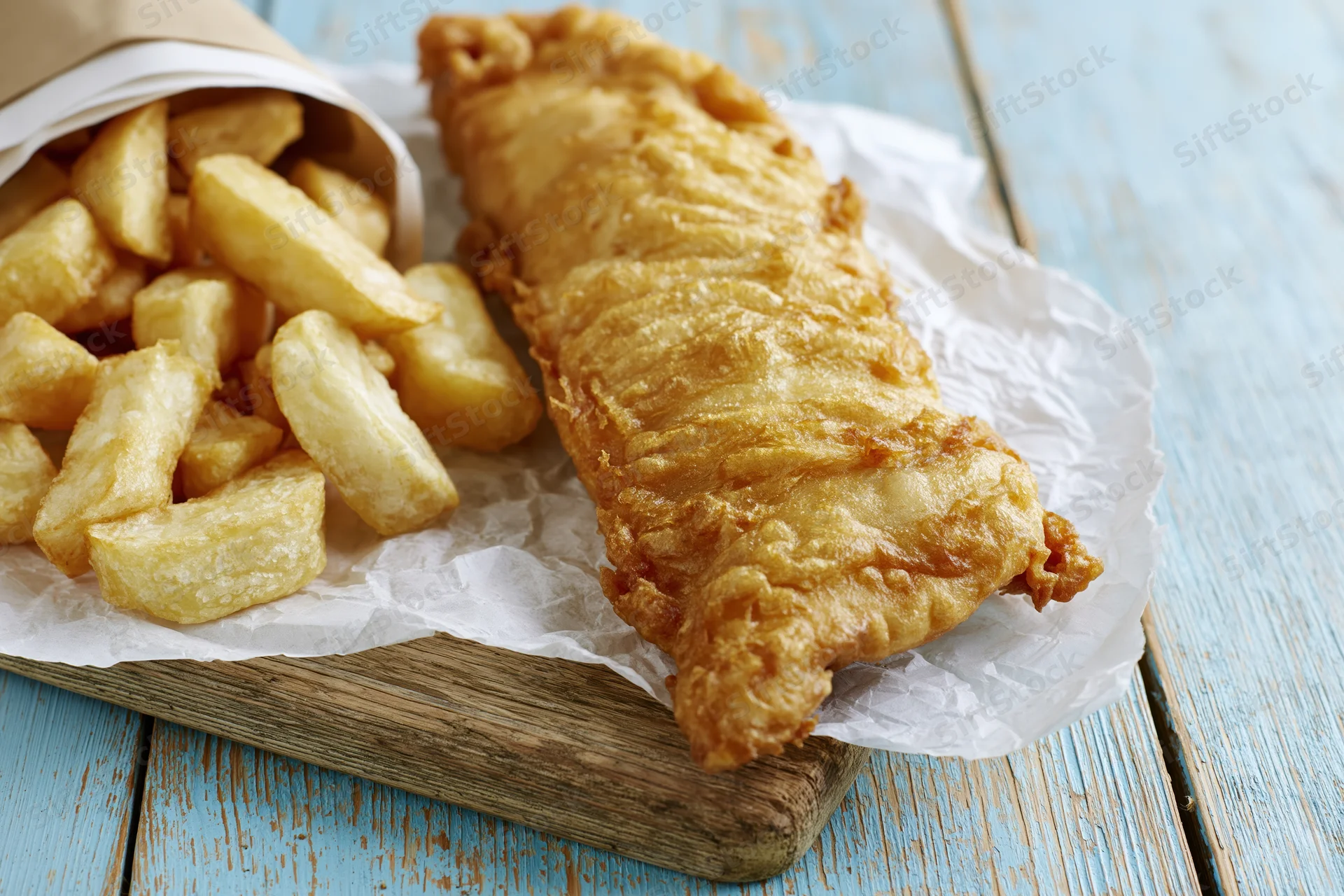 Golden battered fish fillet with thick-cut chips on crumpled paper atop a wooden board