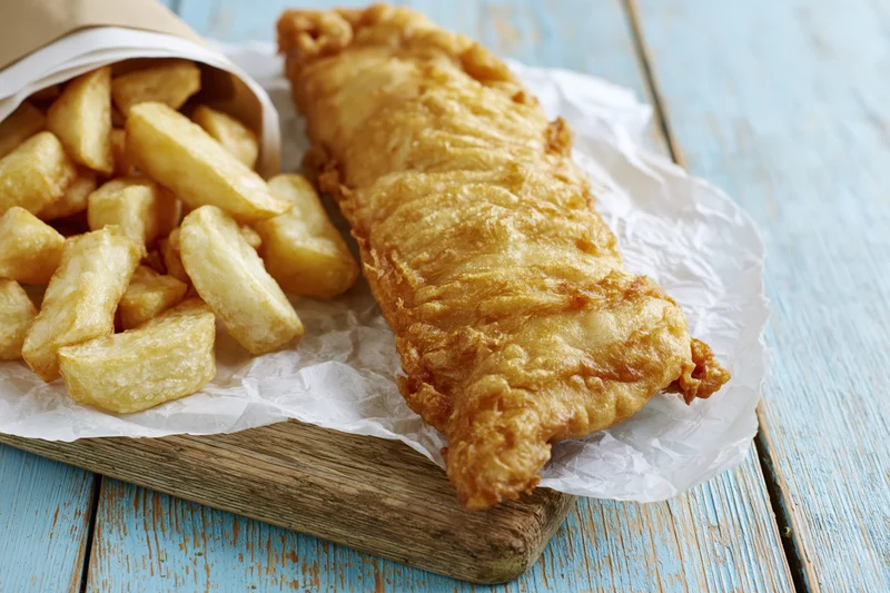 Golden battered fish fillet with thick-cut chips on crumpled paper atop a wooden board