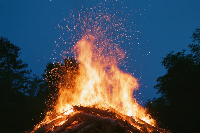 Large outdoor campfire burning intensely at night with sparks rising against dark sky and trees
