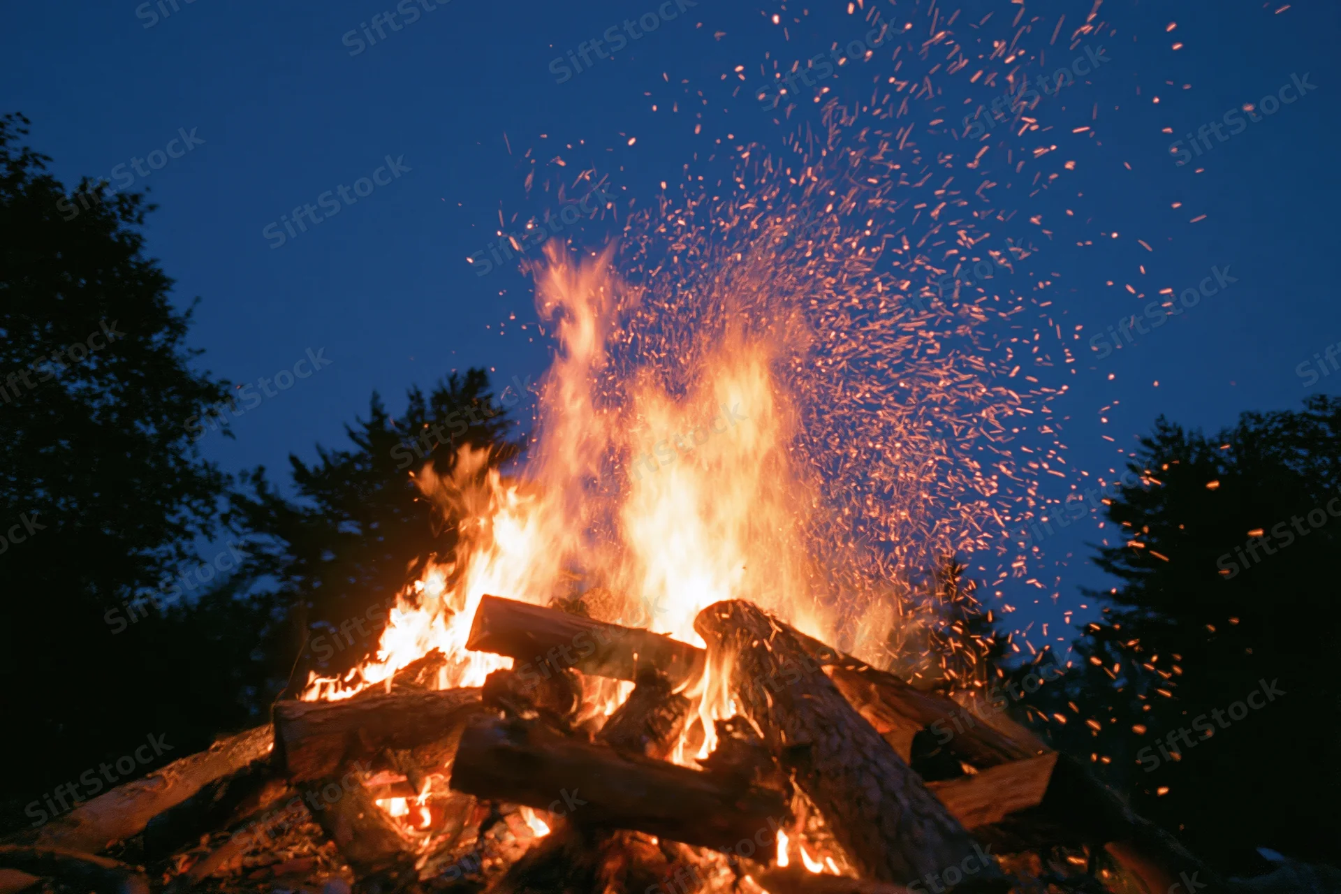 Large campfire burning intensely at night with sparks flying upward against dark sky and trees