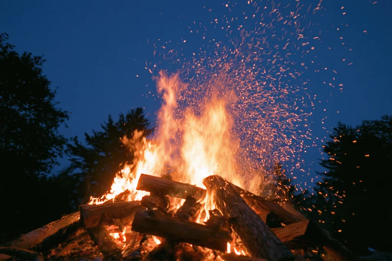 Large campfire burning intensely at night with sparks flying upward against dark sky and trees