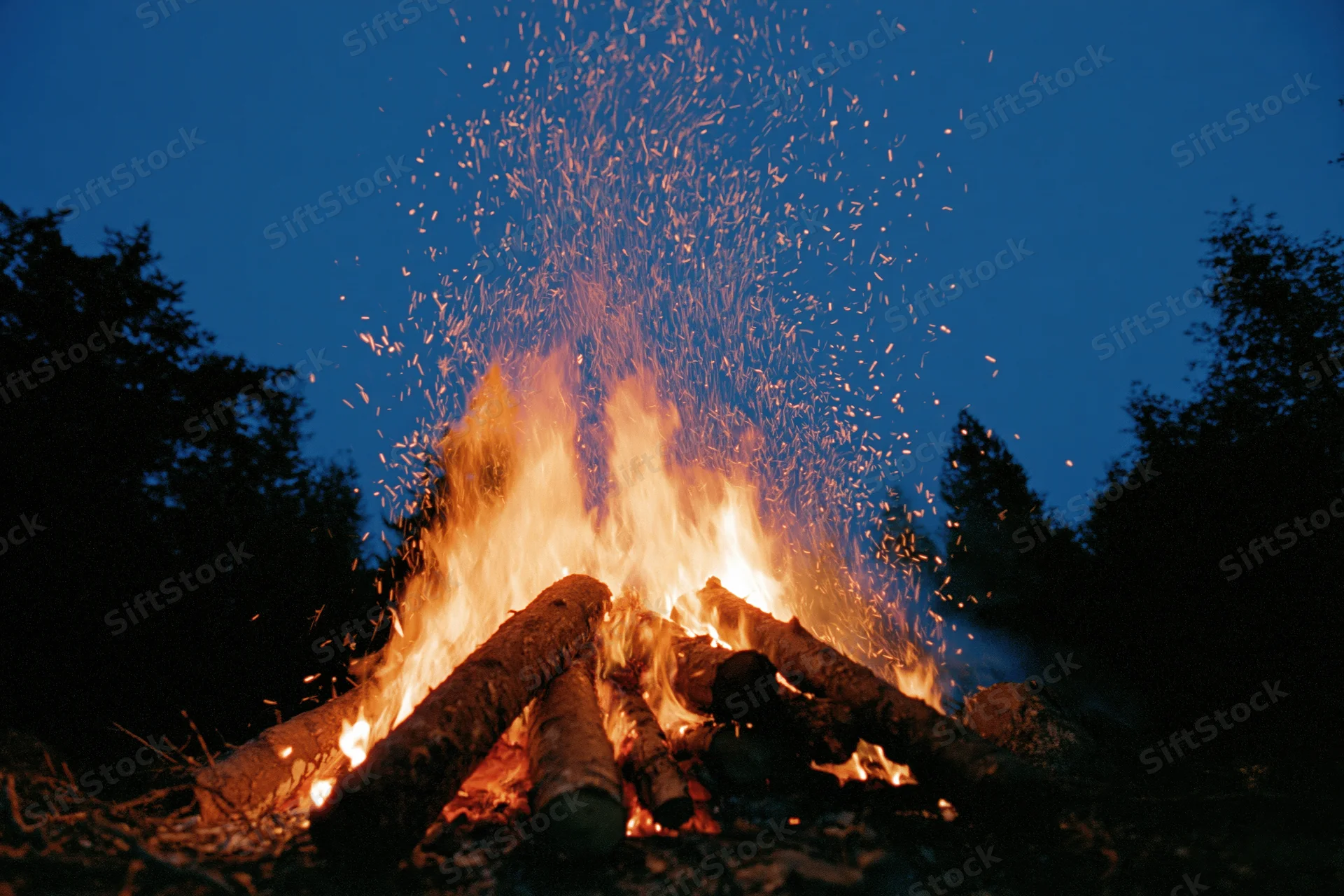 Large campfire burning intensely at night in a forest clearing with sparks rising into the dark blue sky