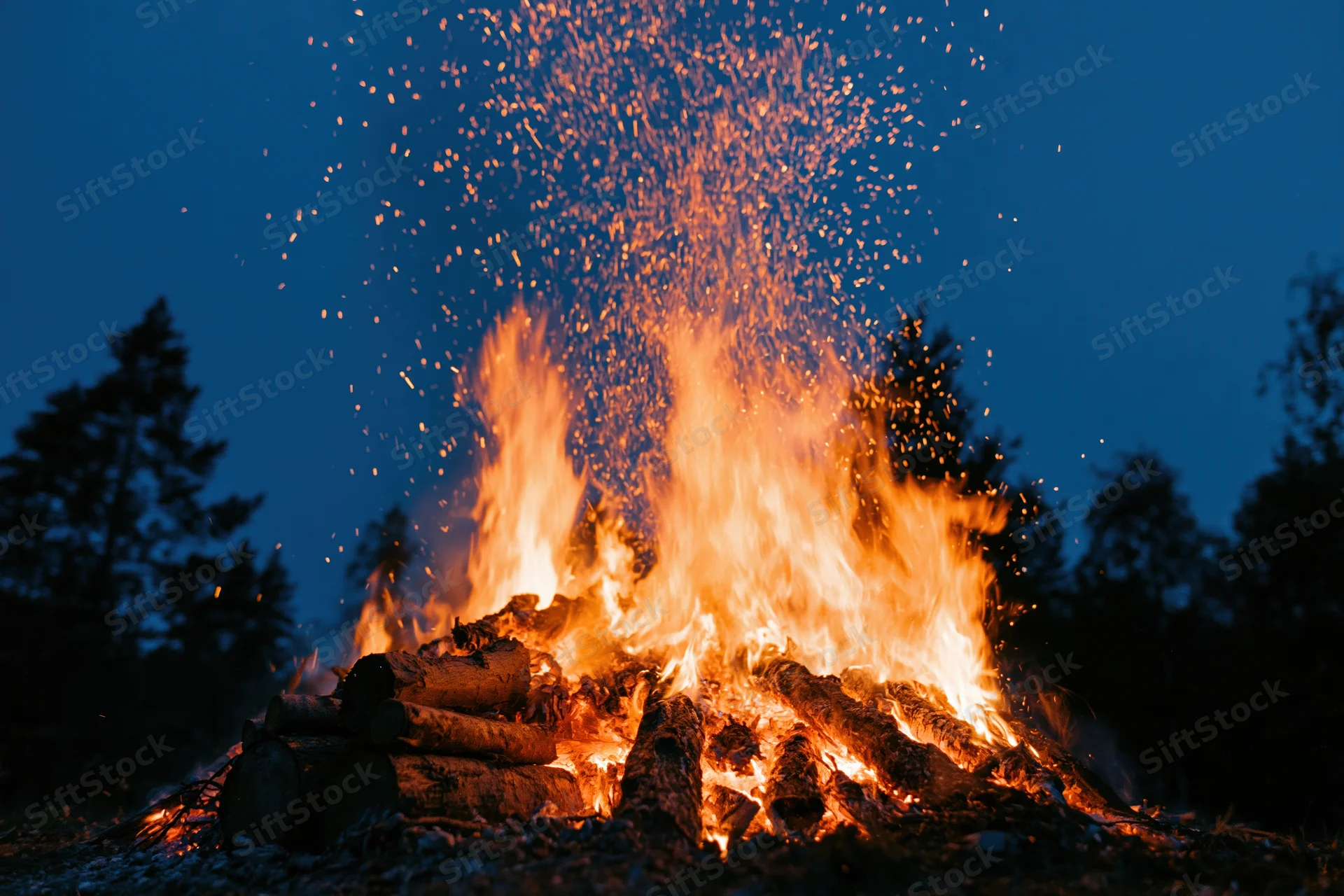 Large campfire burning intensely at night with sparks rising above logs in a forest setting