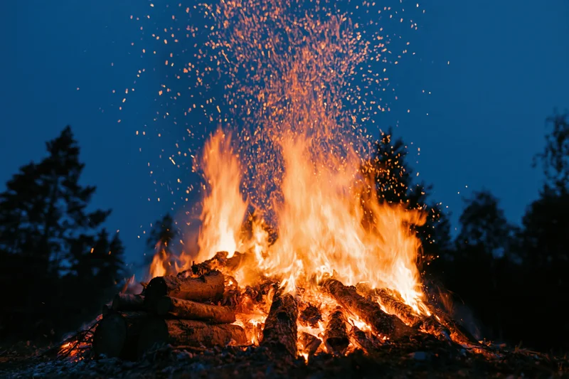 Large campfire burning intensely at night with sparks rising above logs in a forest setting