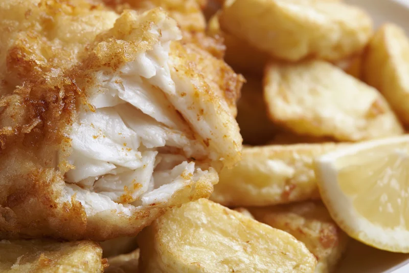 Battered cod fillet broken open to reveal flaky white interior served with chips and lemon slice on plate
