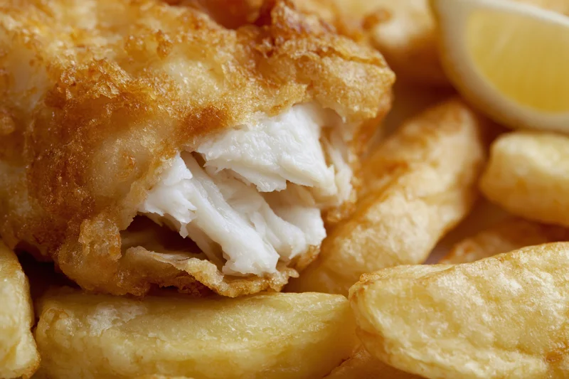 Battered cod fillet broken open to reveal flaky white fish meat served with golden fries and lemon wedge