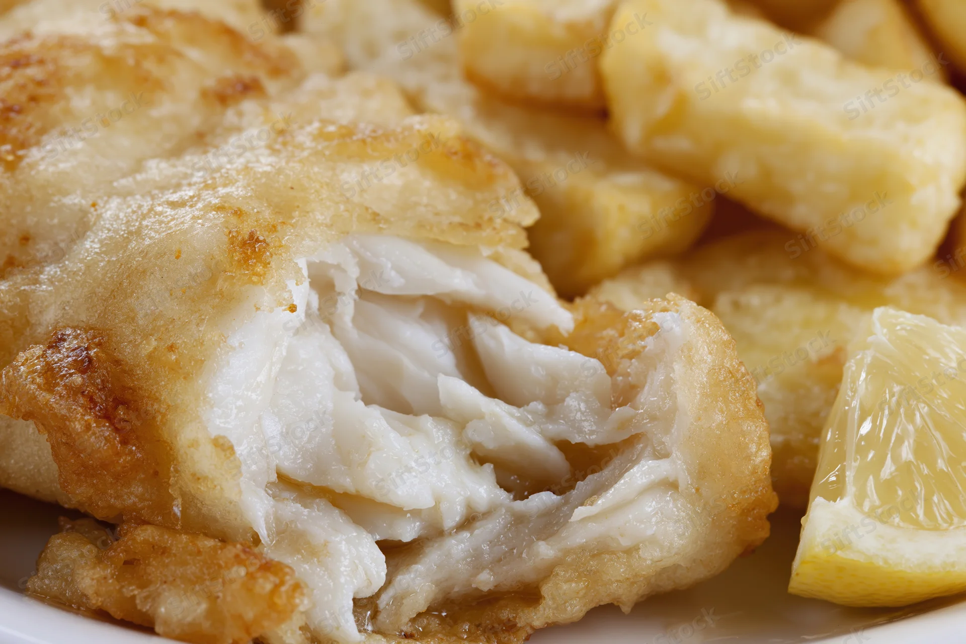 Battered cod fillet broken open to reveal flaky white interior on a plate with lemon wedge and fries