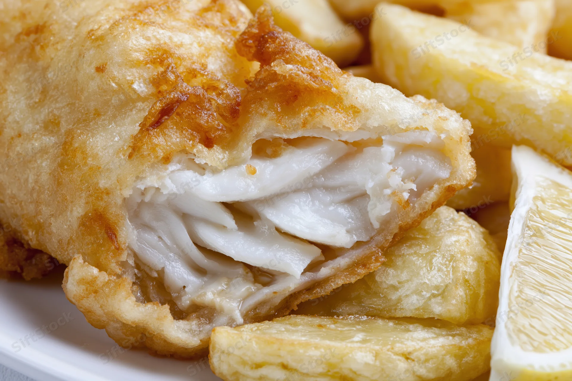 Battered cod fillet broken open to reveal flaky white fish on a plate with chips and lemon wedge
