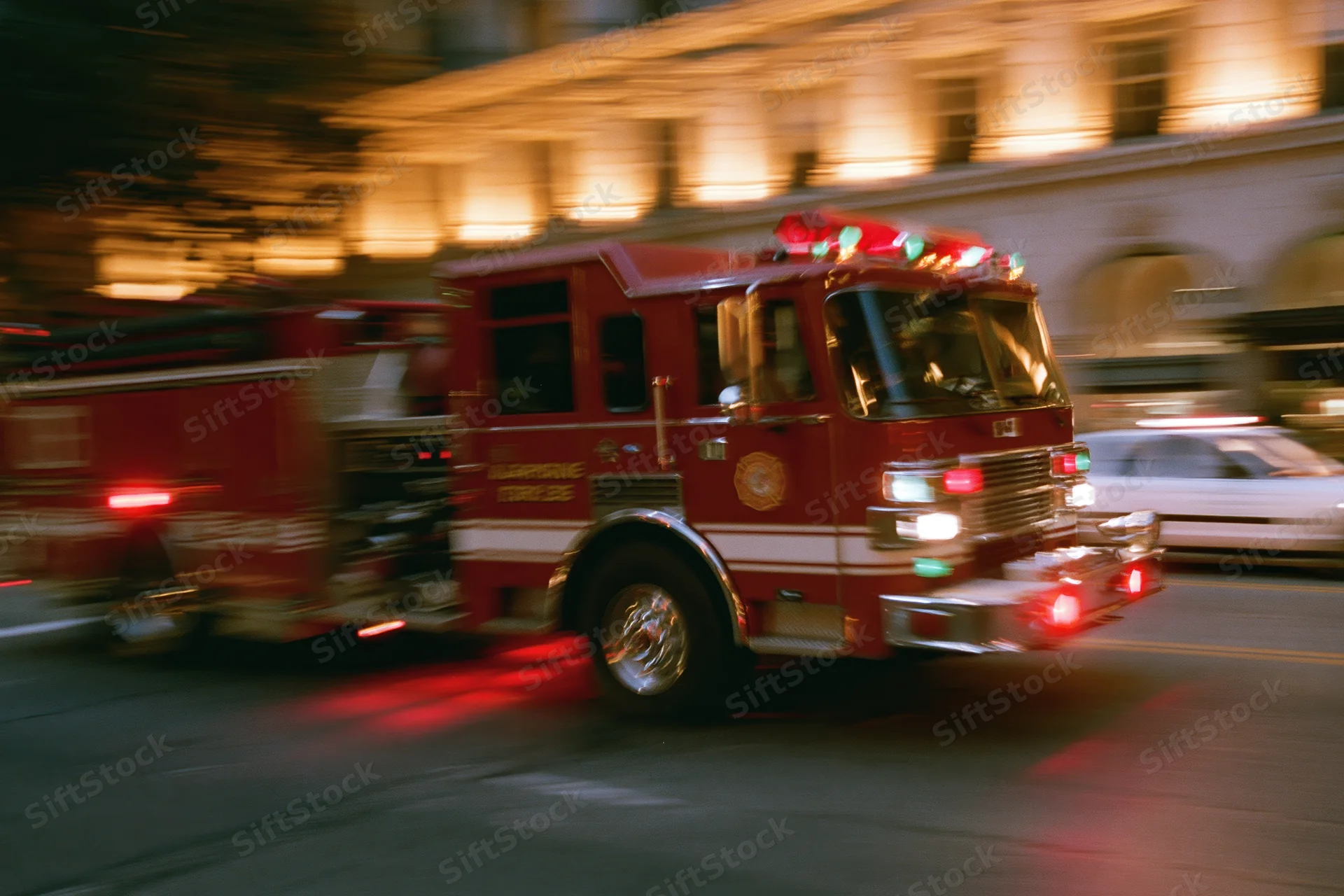 Red fire truck speeding through downtown city intersection at night with flashing lights and motion blur