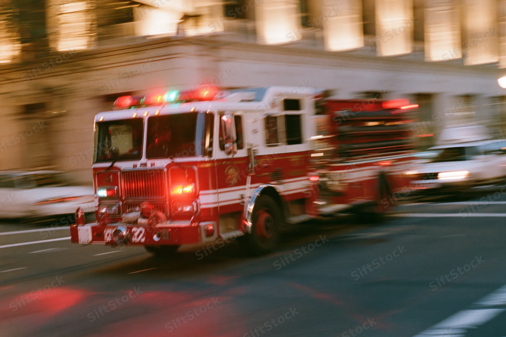 Red fire truck speeding through downtown city intersection at night with flashing lights and motion blur