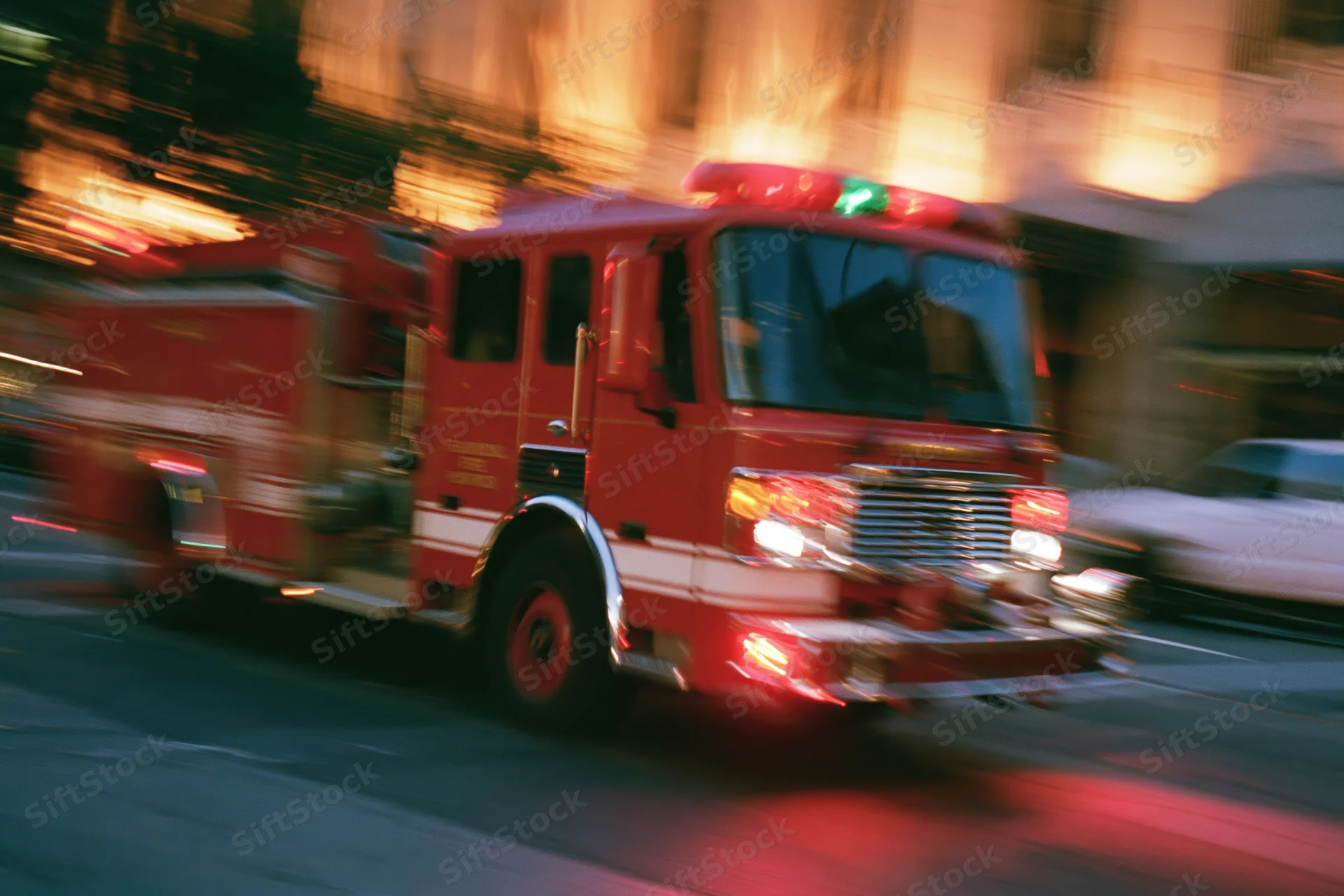 Red fire truck speeding through downtown city intersection at night with flashing lights and motion blur