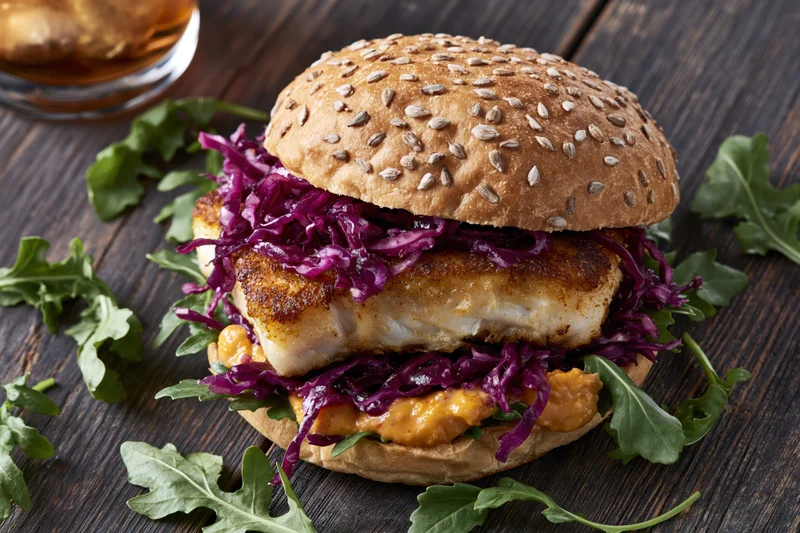 Spiced fish sandwich on a seeded bun with purple cabbage and arugula on dark wood table