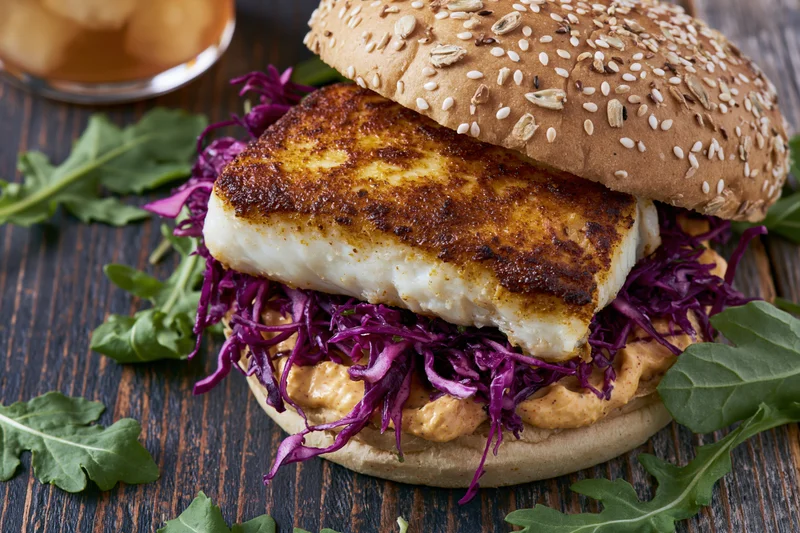 Grilled fish fillet sandwich on a seeded bun with purple cabbage slaw and arugula on a dark wooden table