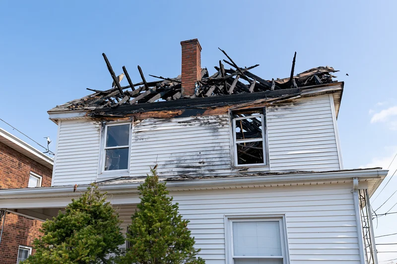 White clapboard house with burned roof and soot-stained siding under clear blue sky