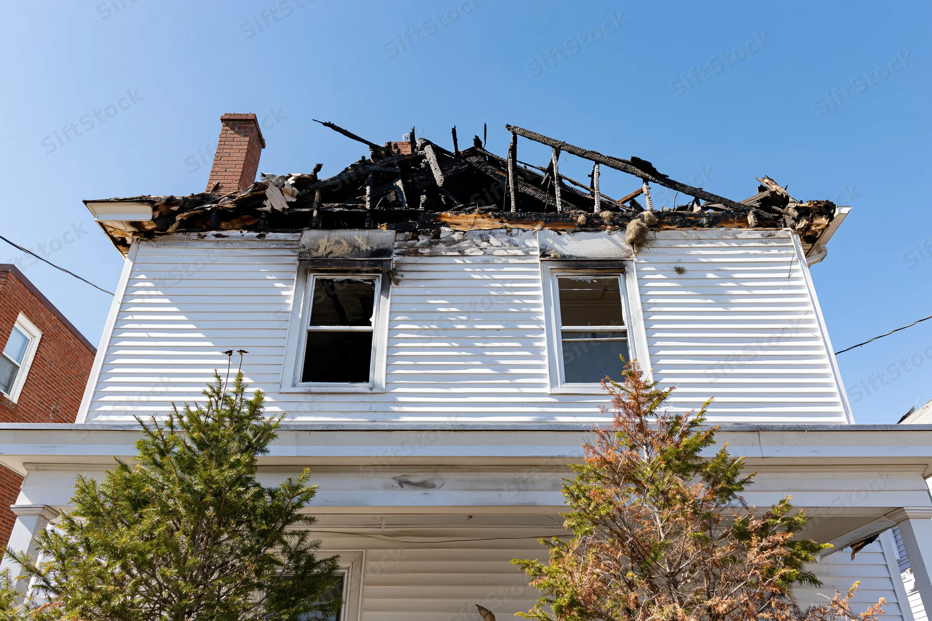 Burned two-story white clapboard house with collapsed roof and charred wood under clear blue sky