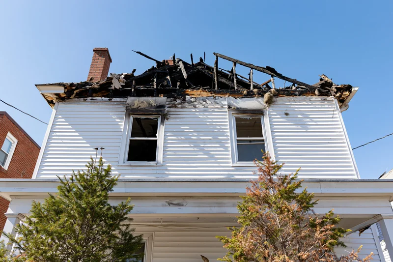 Burned two-story white clapboard house with collapsed roof and charred wood under clear blue sky