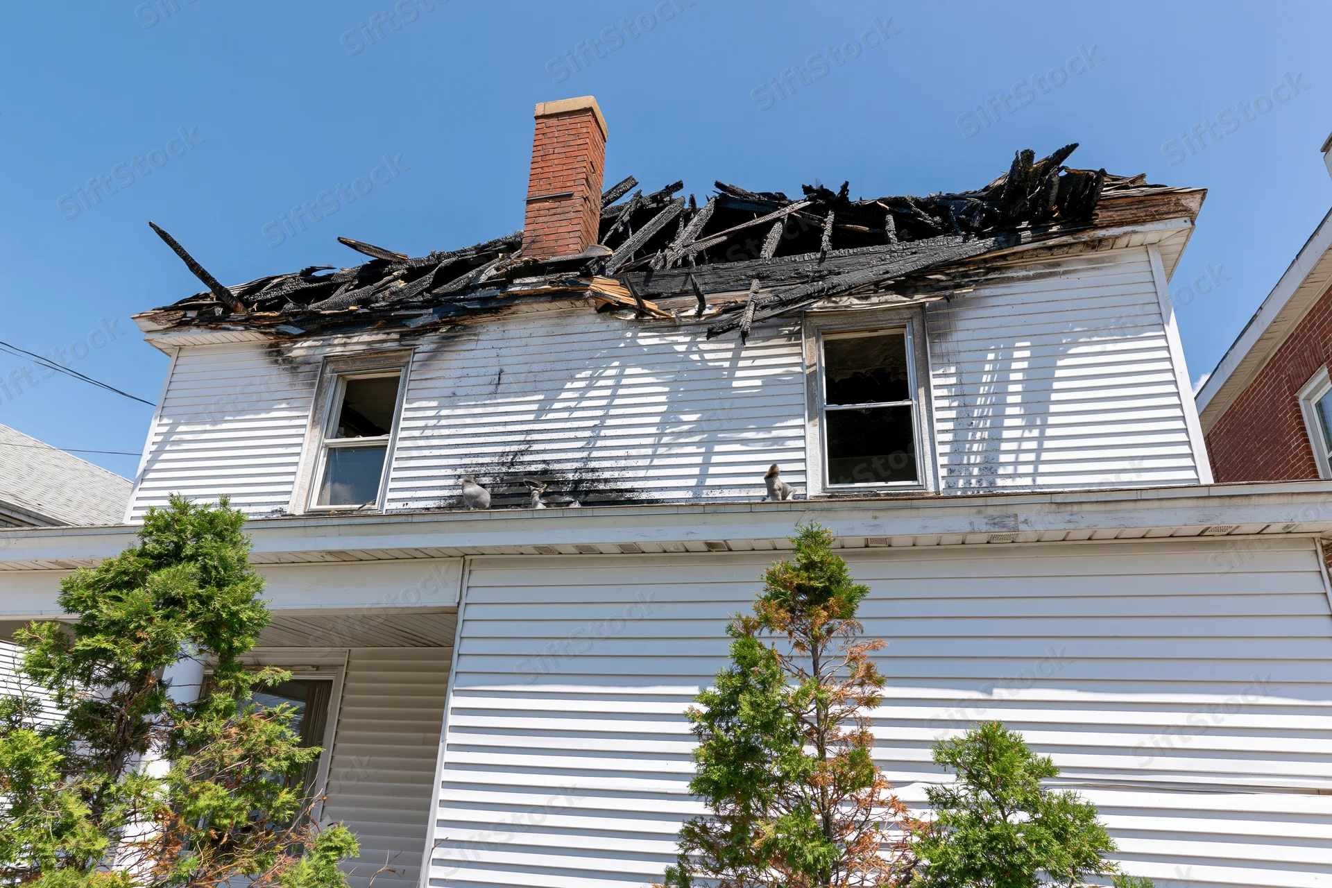 Two-story white clapboard house with burned roof and broken windows under clear blue sky