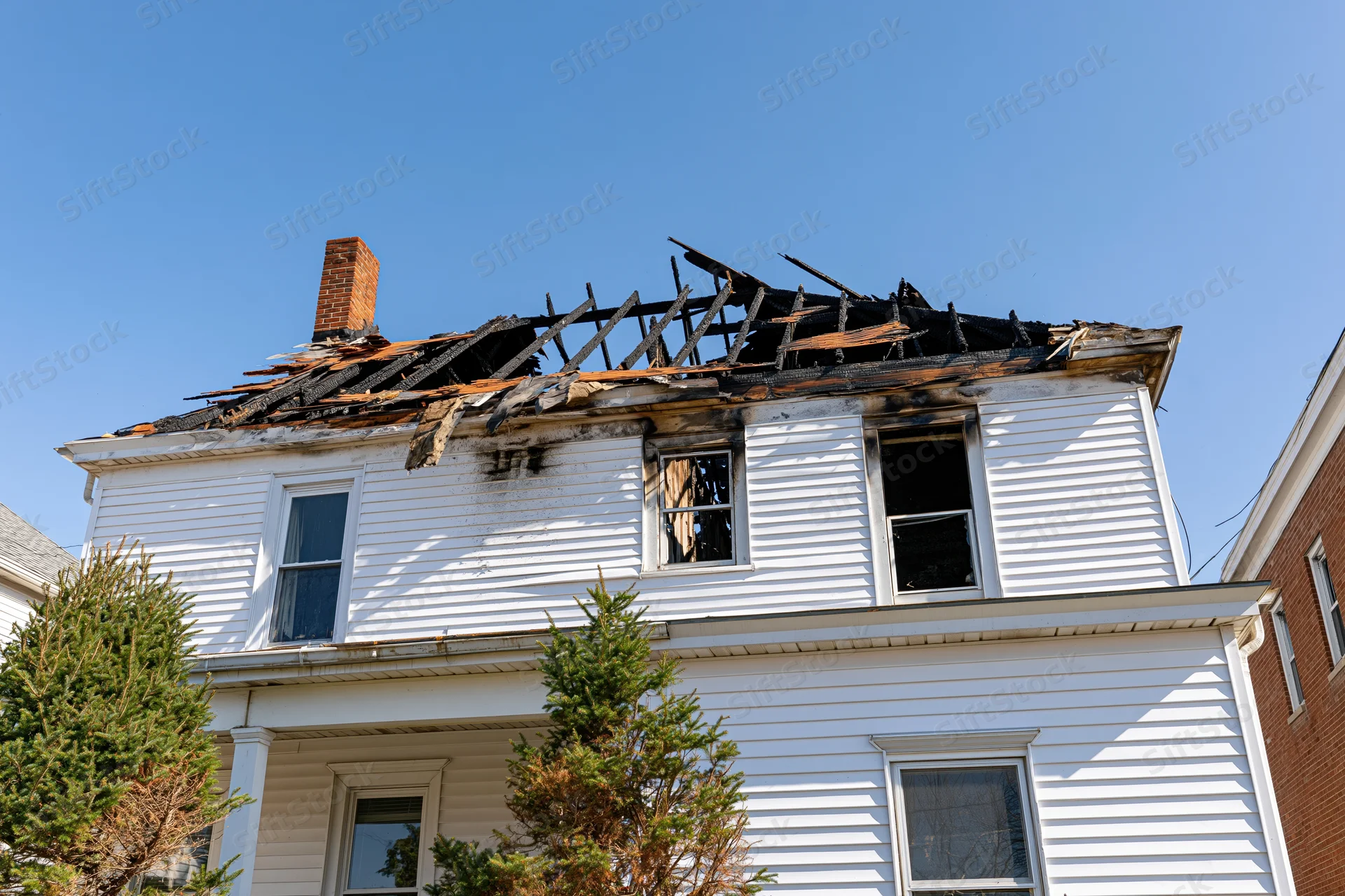 Two-story white clapboard house with burned roof and broken windows under clear blue sky