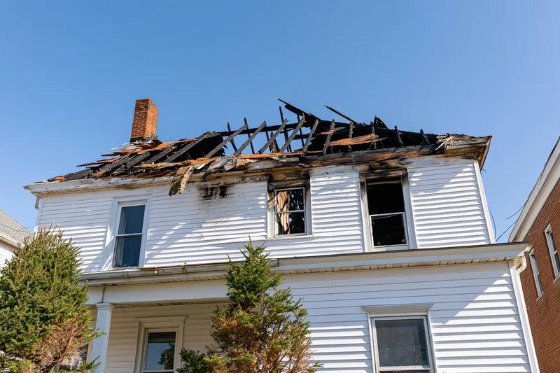 Two-story white clapboard house with burned roof and broken windows under clear blue sky