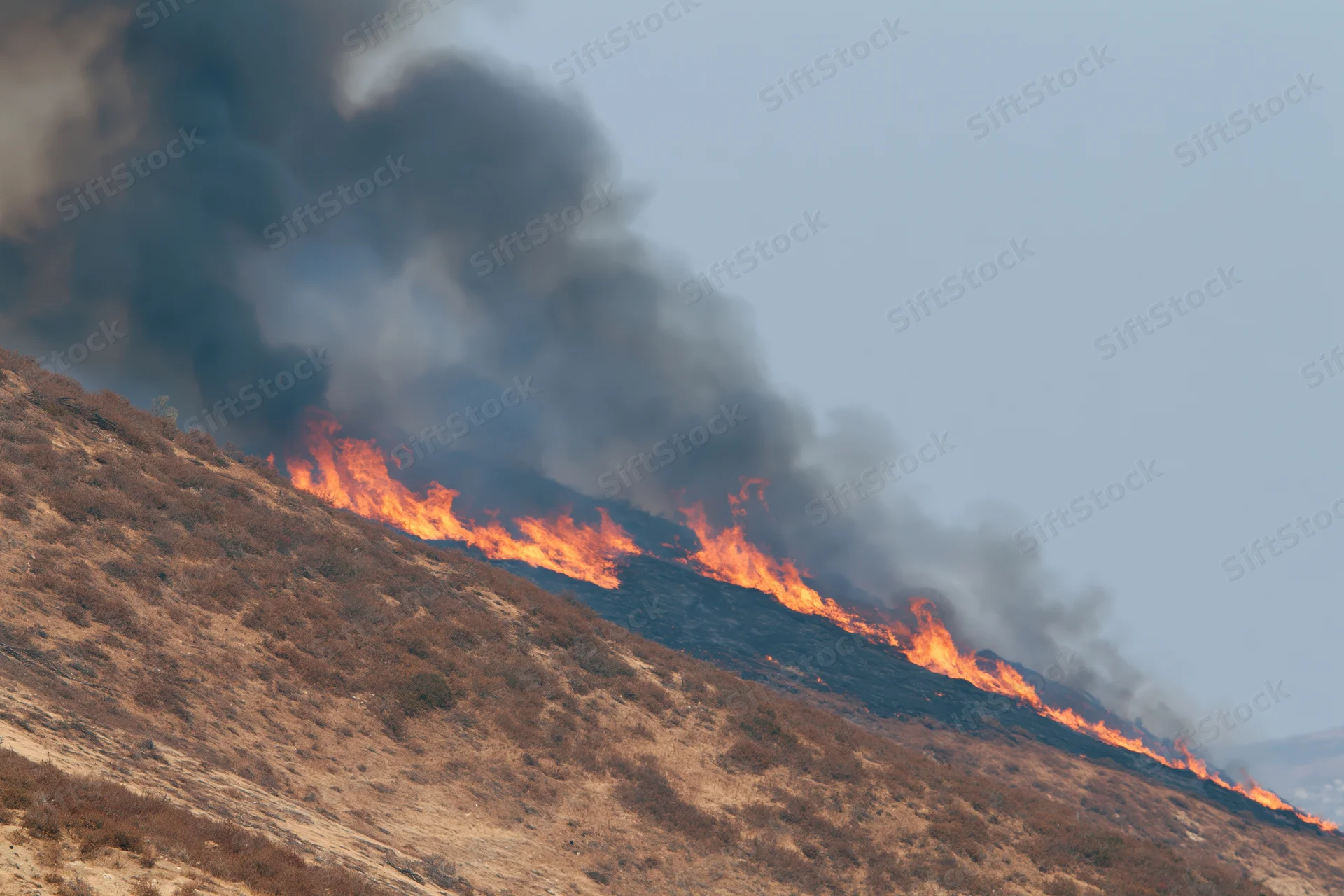 Wildfire burning along a dry hillside ridge with thick smoke rising into the sky