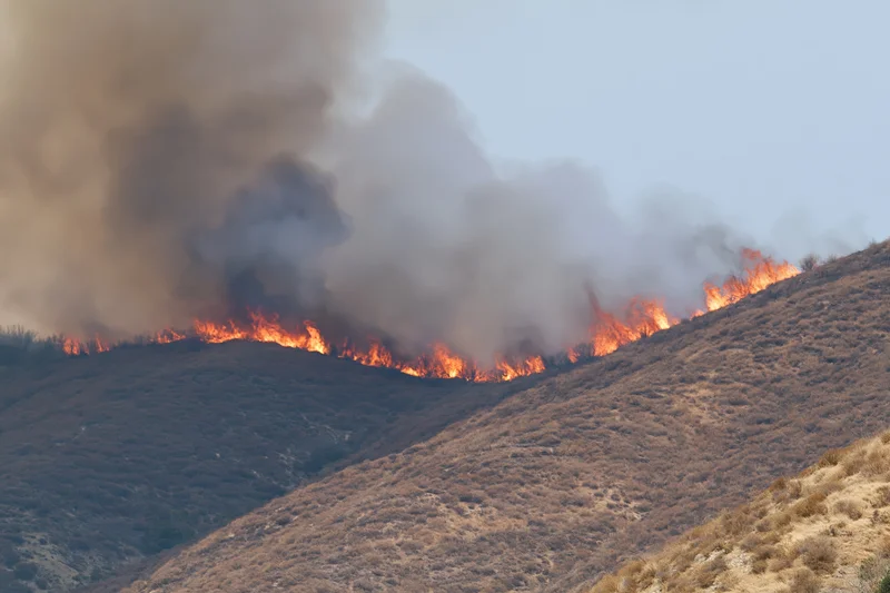 Wildfire burning along a dry hillside ridge with thick smoke rising into the sky