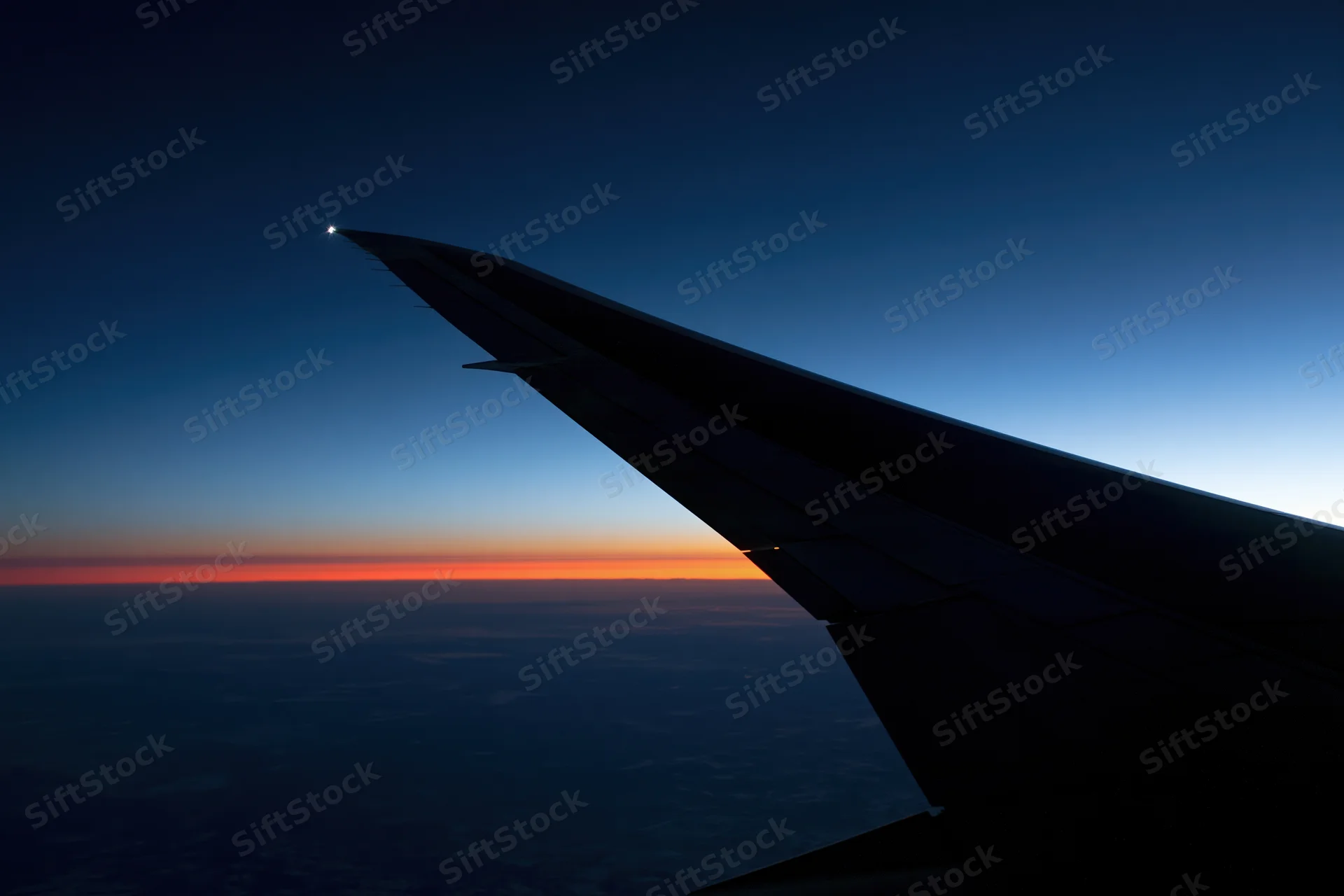 Airplane wing silhouetted against twilight horizon with orange sky and clouds below