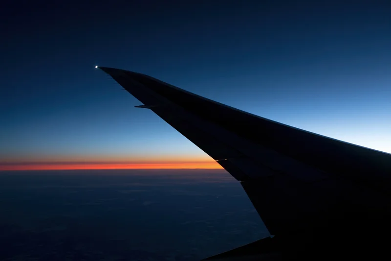 Airplane wing silhouetted against twilight horizon with orange sky and clouds below