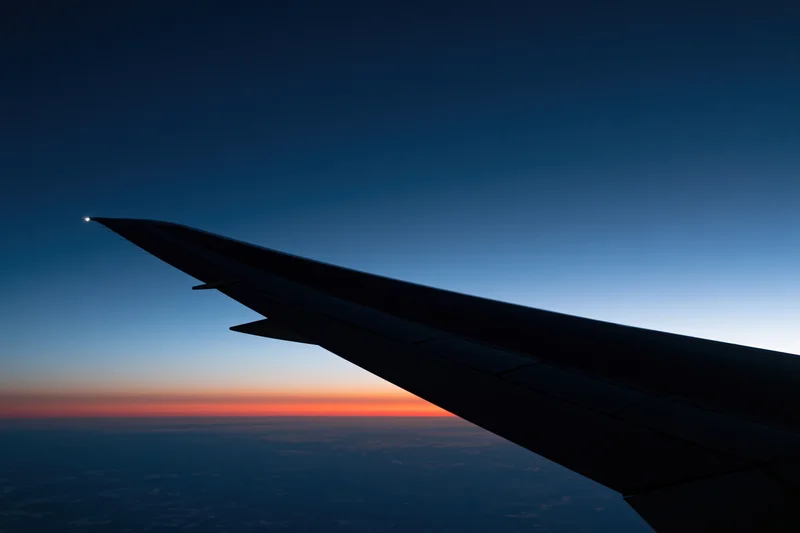 Airplane wing silhouetted against twilight sky with orange horizon and clouds below